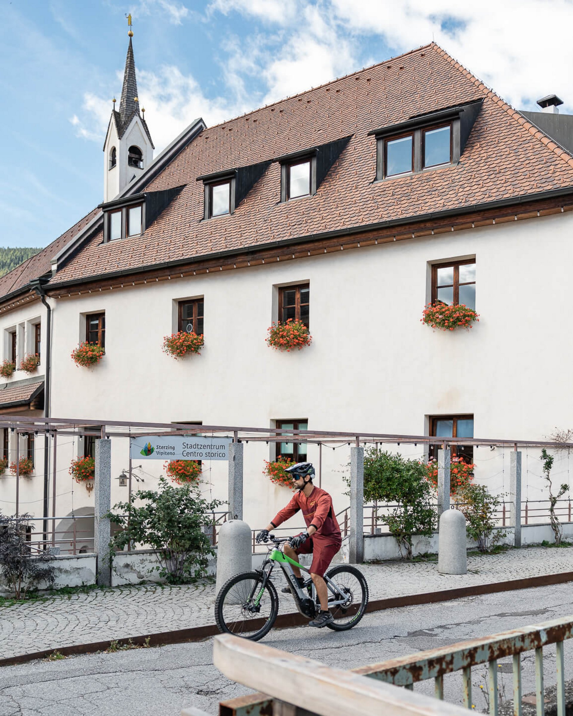 Mann fährt Fahrrad vor einem traditionellen Gebäude mit Kirchturm und Blumenfenstern