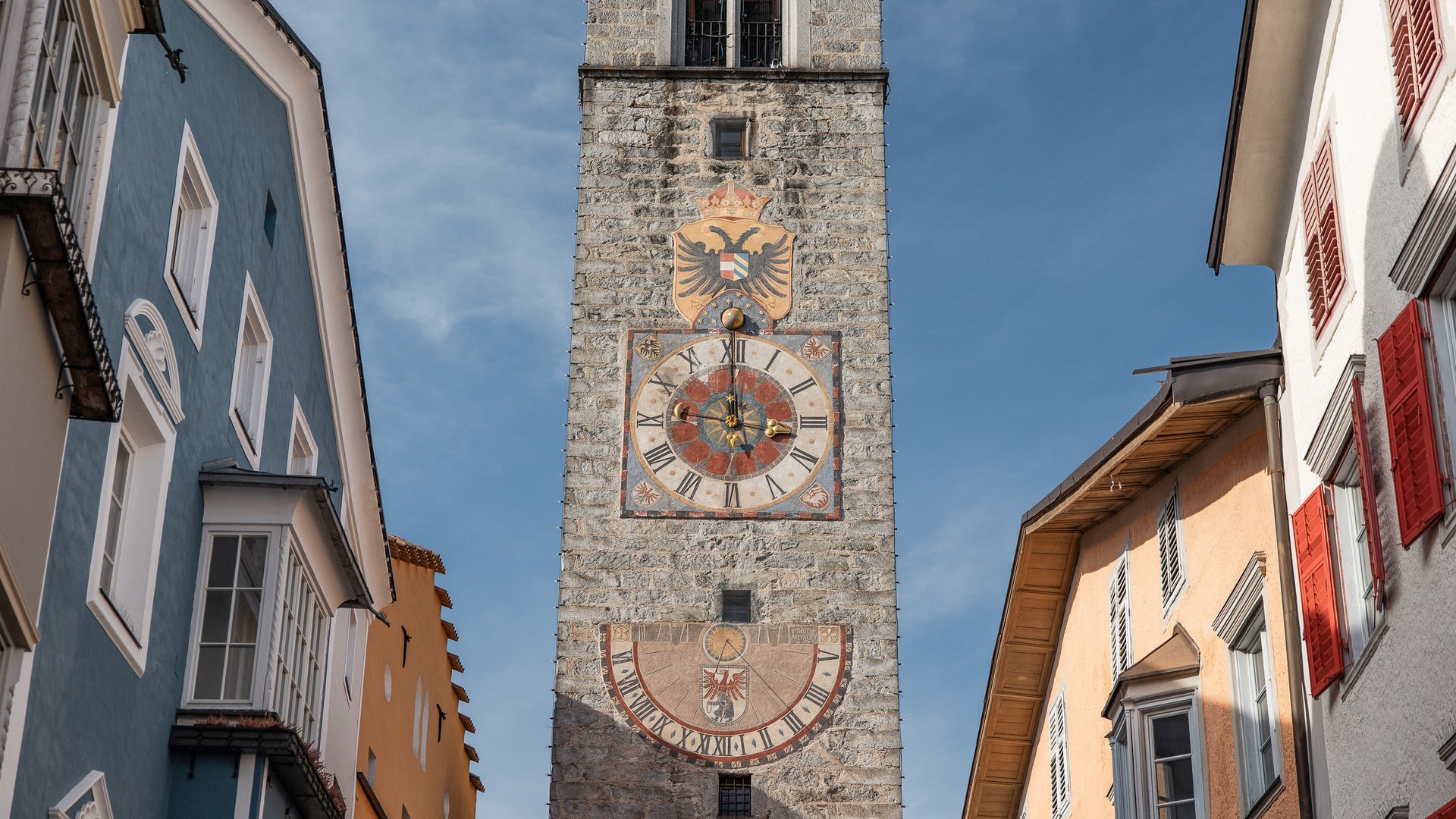 Historischer Uhrenturm aus Stein mit zwei großen Zifferblättern zwischen bunten Häusern