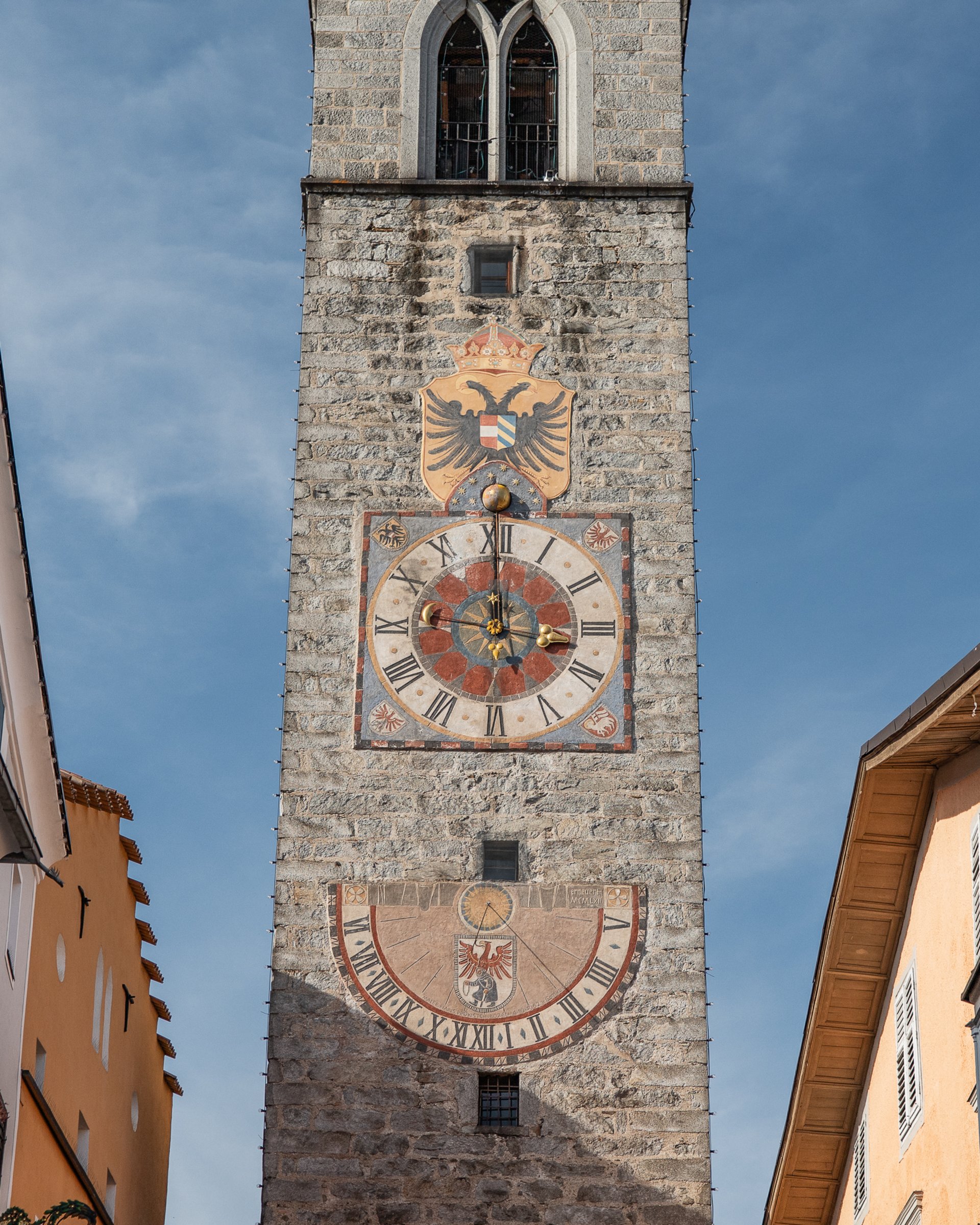 Historischer Uhrenturm aus Stein mit zwei großen Zifferblättern zwischen bunten Häusern
