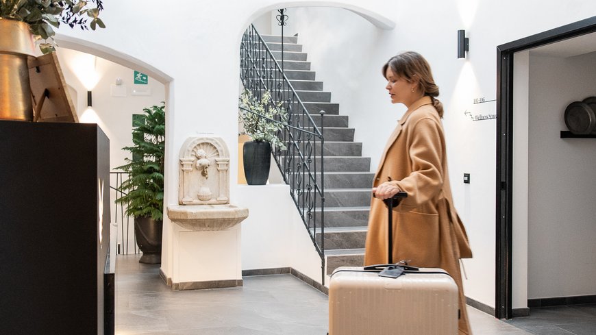 Woman with suitcase walking through bright hotel hallway with stairs in background