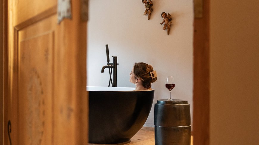 Frau entspannt in schwarzer Badewanne, mit Weinglas auf Hocker, gesehen durch Tür