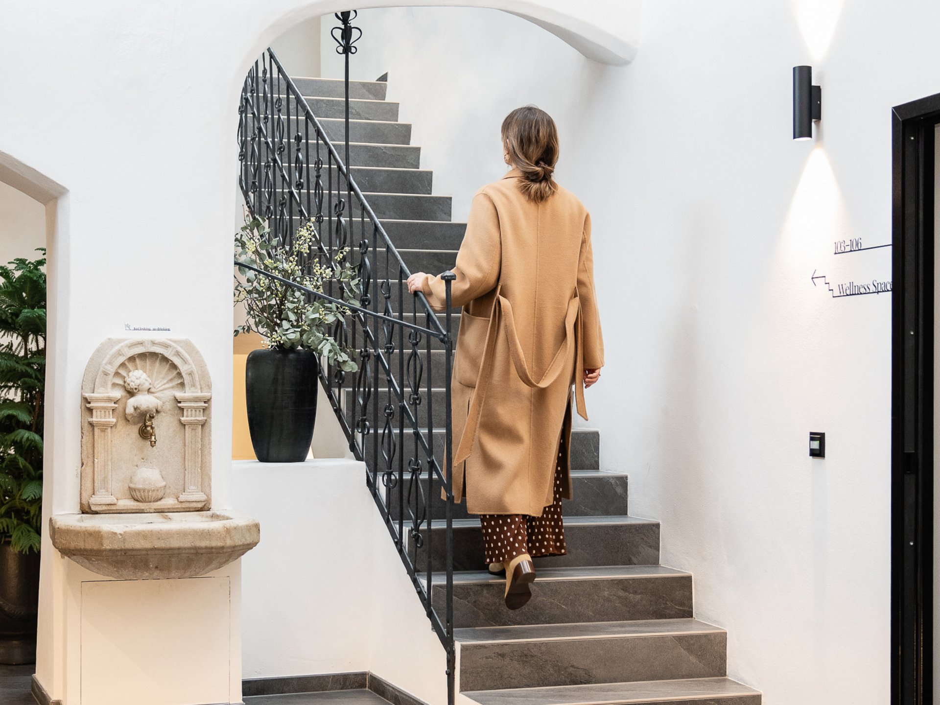 Frau in braunem Mantel steigt Treppe in modernen Innenraum hinauf