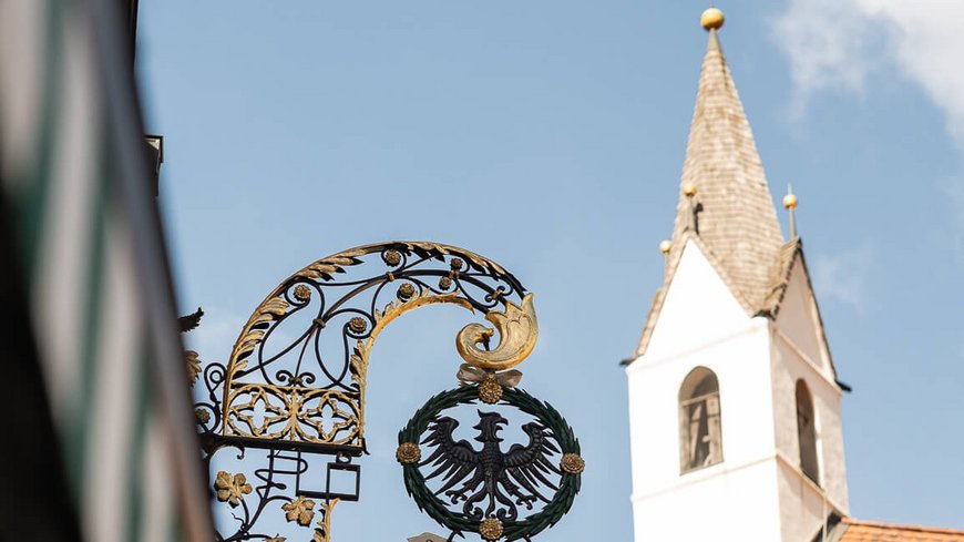 Die perfekte Location für euer Apartment in Sterzing Schild mit schwarzem Adler und Kirche mit Turm bei blauem Himmel