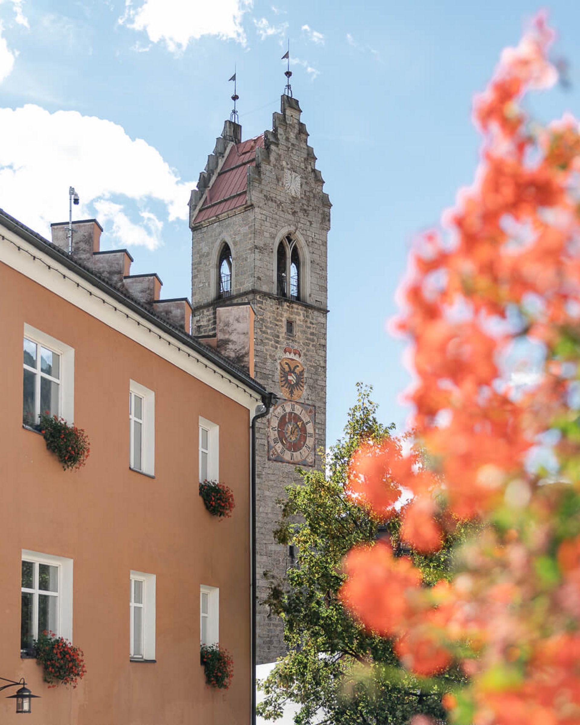 Turm mit Uhr an einem historischen Gebäude neben einem orangen Haus mit Blumen an Fenstern