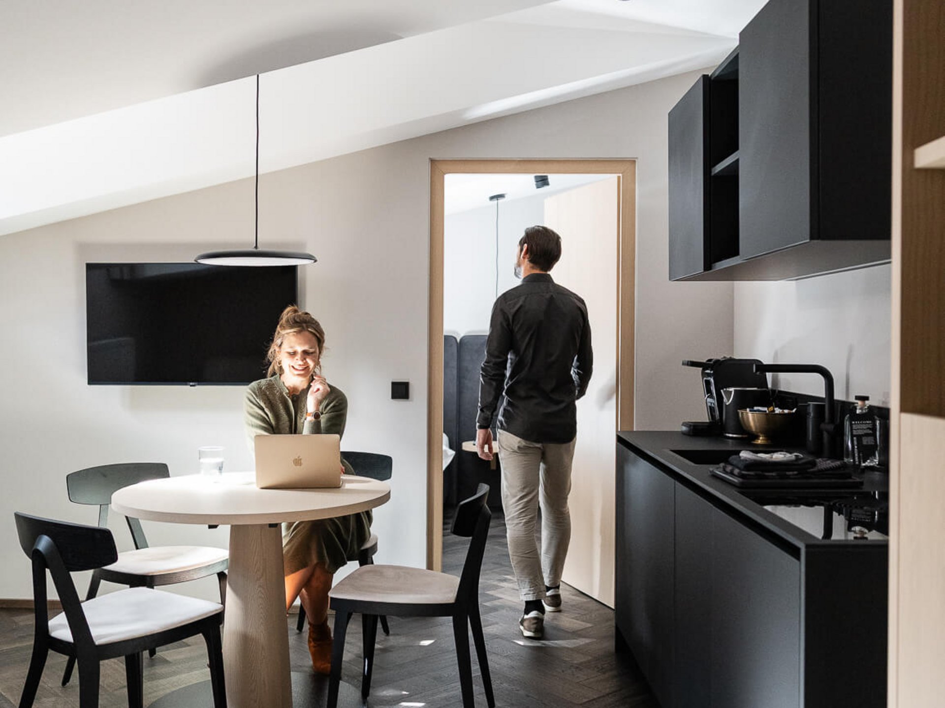 Frau arbeitet am Laptop am Küchentisch, Mann geht durch Tür im modernen Apartment