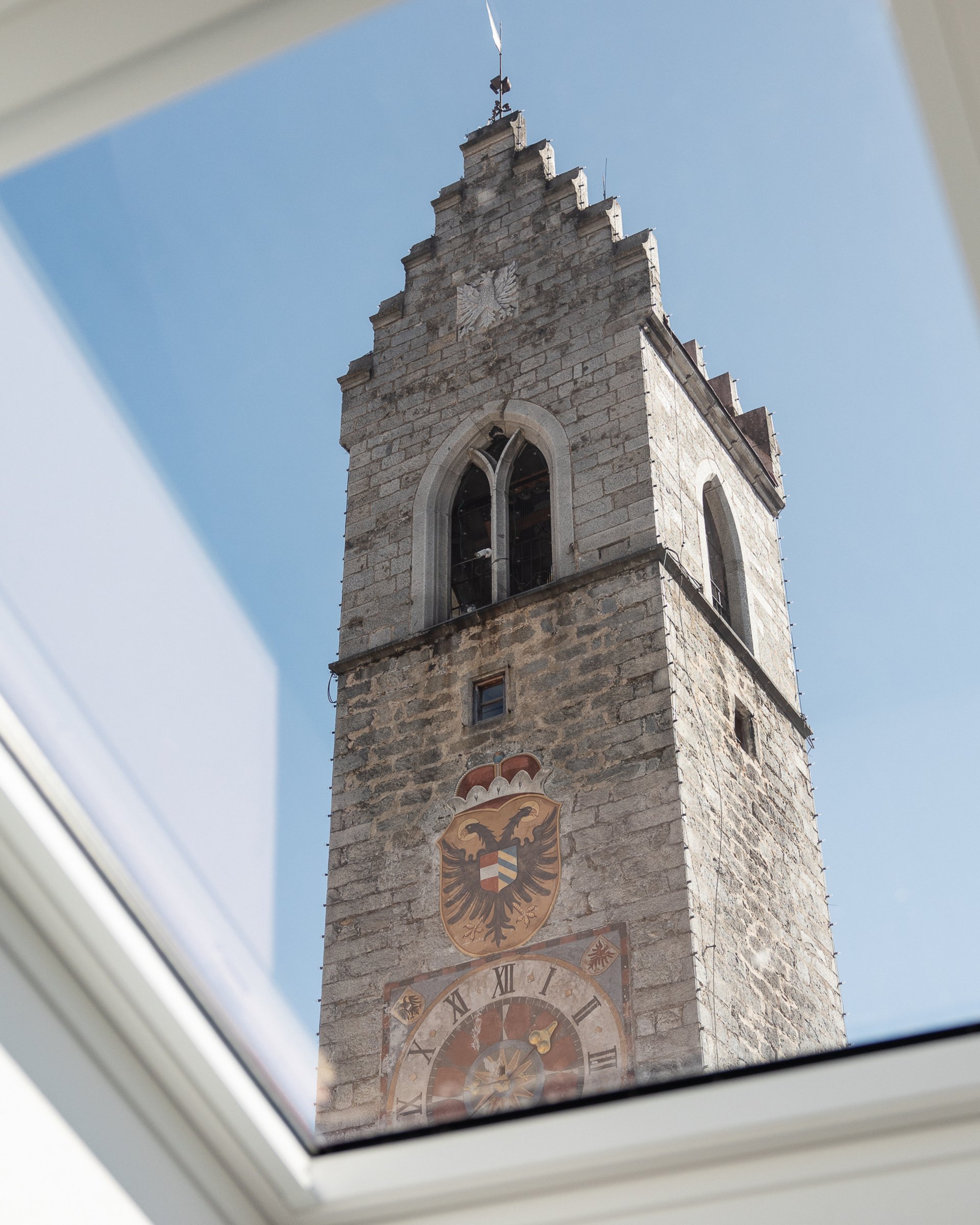 Turm mit Uhr und Wappen vor blauem Himmel, durch ein Fenster gesehen
