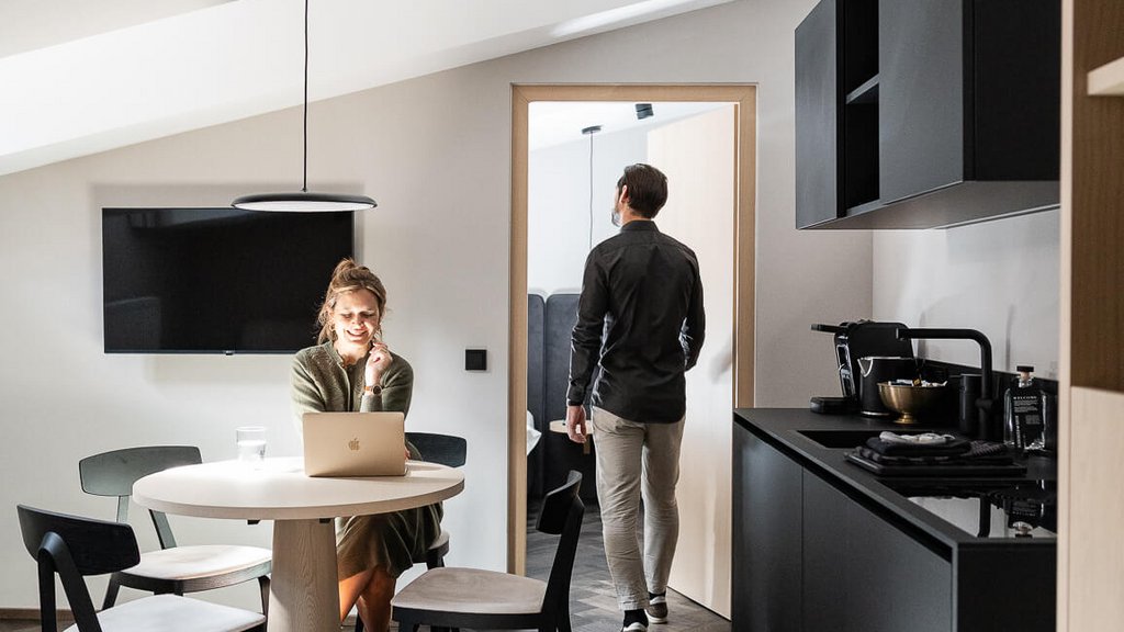 Frau arbeitet mit Laptop am Tisch, Mann geht durch Zimmertür in moderner Wohnung