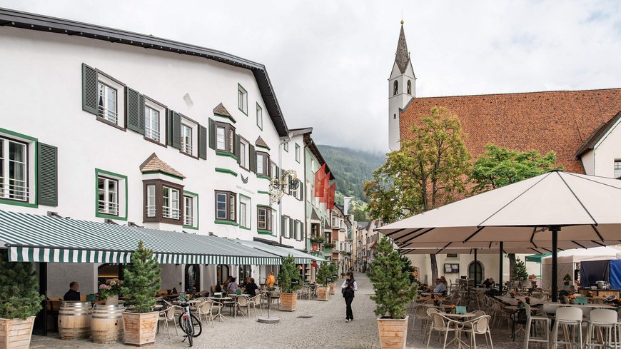 Die perfekte Location für euer Apartment in Sterzing Straßencafé in einem malerischen europäischen Dorf mit Kirche und Bergen im Hintergrund