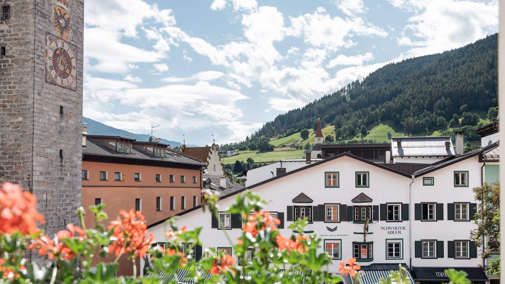 Die perfekte Location für euer Apartment in Sterzing Altstadt mit Turmuhr, Blumen und Bergen im Hintergrund an einem sonnigen Tag