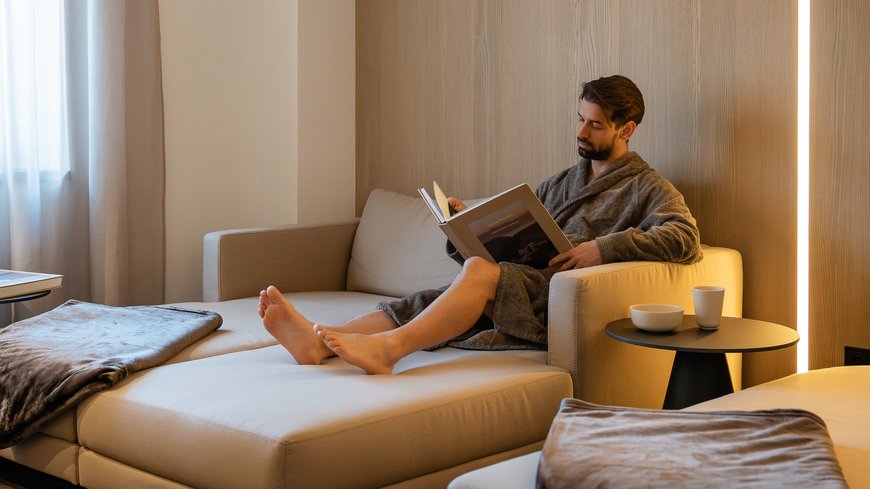 Benessere all’Aquila Nera Uomo in accappatoio che legge rilassato su un divano beige in un soggiorno moderno.