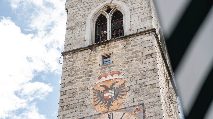 Die perfekte Location für euer Apartment in Sterzing Historischer Uhrenturm aus Stein mit Wappen bei blauem Himmel