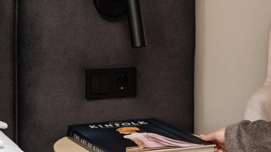 A person reaching for a book on a round bedside table next to a bed.
