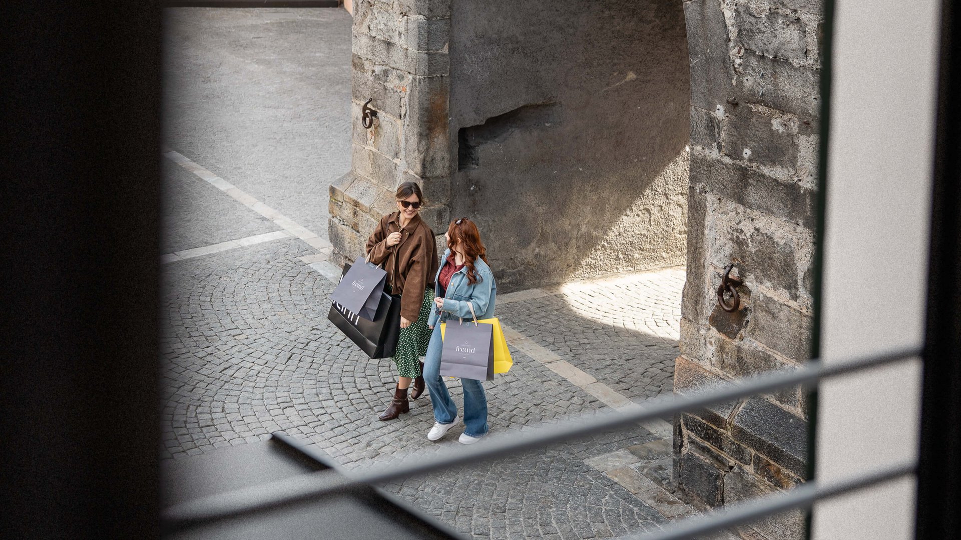 Zwei Frauen mit Einkaufstaschen gehen auf gepflasterter Straße neben einem alten Steinbogen