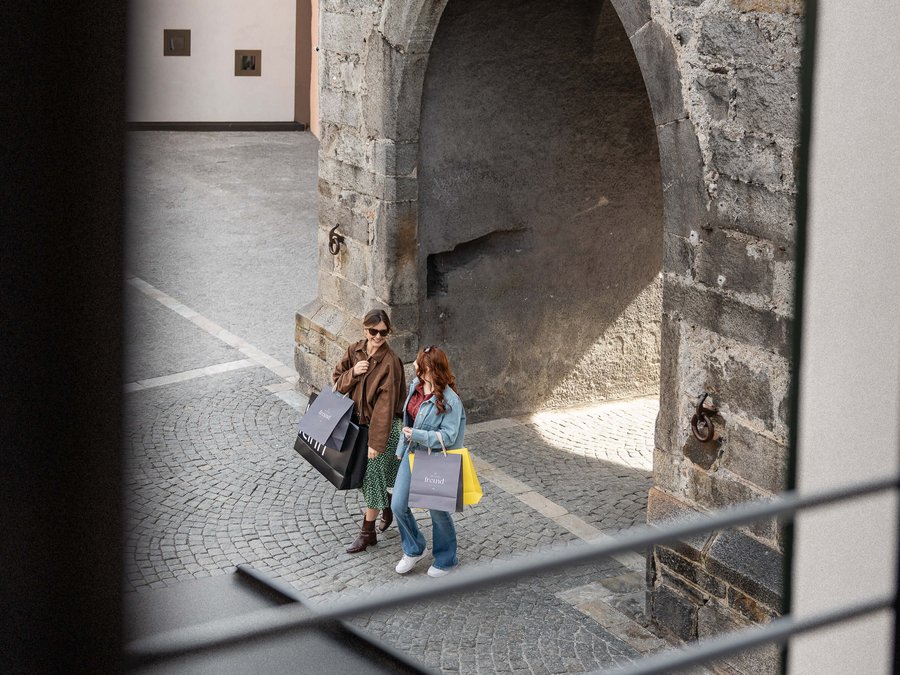 Aquila Nera: la storia del nostro albergo a Vipiteno Due donne con borse della spesa camminano su strada acciottolata vicino a un antico arco di pietra
