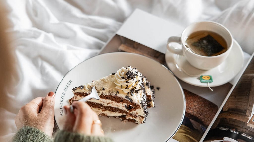 Hand hält Teller mit Stück Kuchen neben Tasse Tee auf Bett mit Magazin
