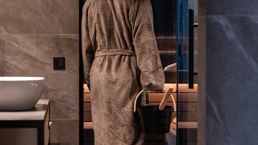 Person in bathrobe entering sauna holding bucket and ladle