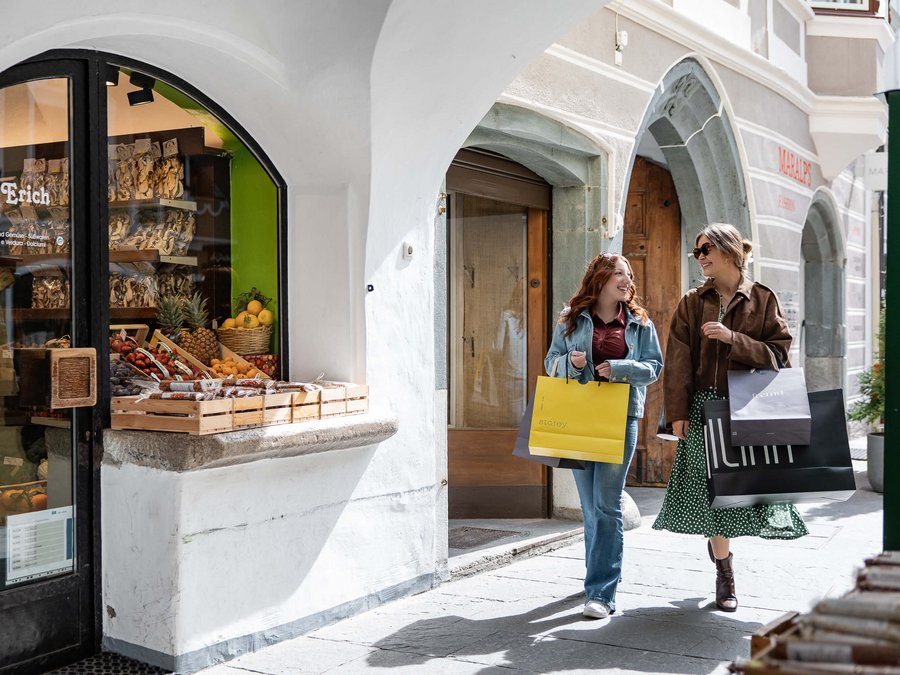 Süße Boutiquen, edle Delikatessengeschäfte Zwei Frauen gehen mit Einkaufstüten durch eine Straße mit Bögen und Geschäften