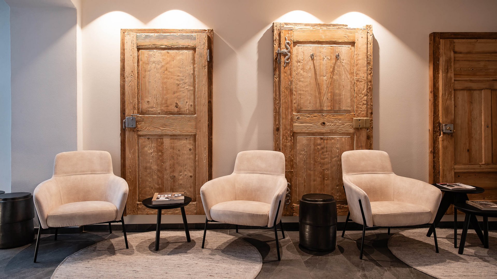 Drei beige Sessel vor drei hellen Holztüren an einer Wand mit runden Teppichen