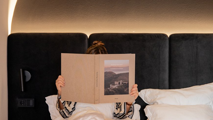 Person liest Buch im Bett mit schwarzem Kopfteil und warmem Lichtbogen