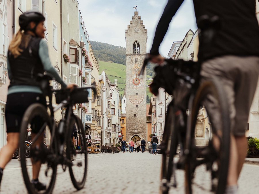 Schwarzer Adler Fragen und Antworten Zwei Radfahrer gehen durch eine Altstadtstraße mit historischem Turm im Hintergrund